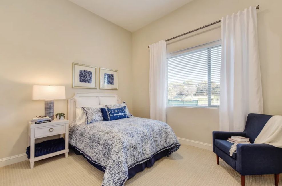 A cozy bedroom featuring a comfortable bed with patterned bedding, a side table with a lamp, and large window with curtains.