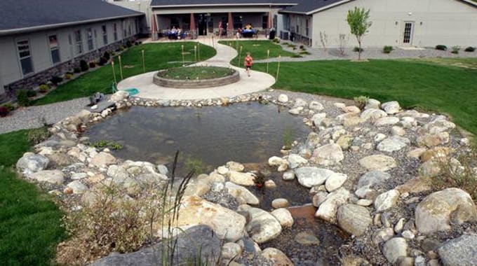 A backyard landscape featuring a pond surrounded by rocks, with a grassy area and a pathway leading to a building.