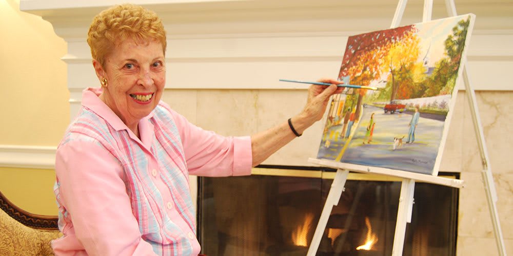 A woman smiling while painting on a canvas at an easel indoors.