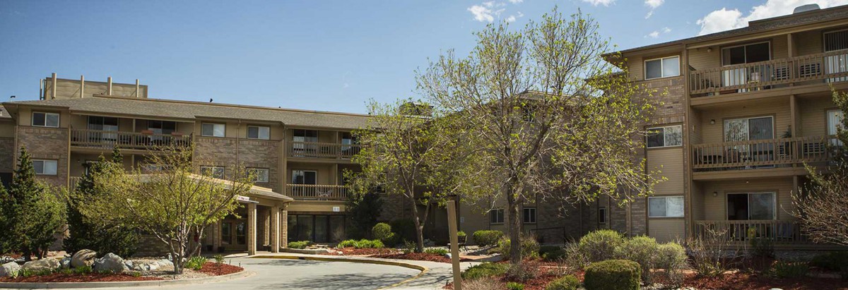 A residential apartment complex surrounded by trees and landscaping under a clear blue sky.
