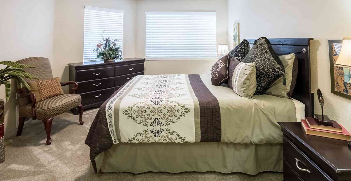 A well-decorated bedroom with a neatly made bed, accent pillows, and a dresser by the window.