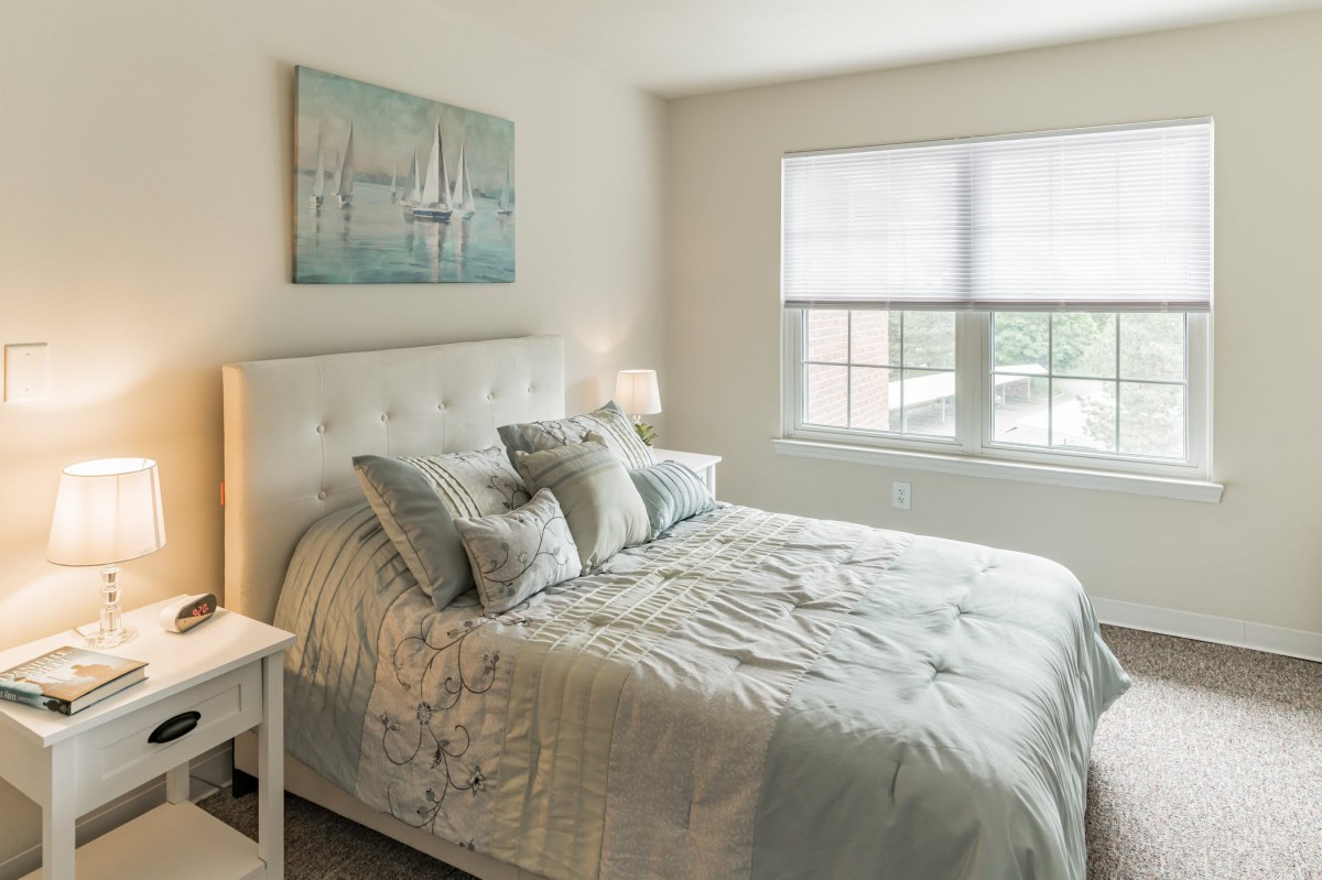 A cozy bedroom with a neatly made bed, adorned with multiple pillows and a bedside lamp. The room features a large window and wall art depicting sailboats.