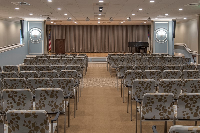 An empty auditorium with rows of chairs facing a stage with closed curtains and a piano.