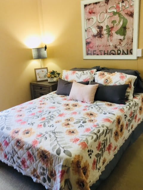 A cozy bedroom with floral bedding and decorative pillows, featuring wall art and a warm lamp.