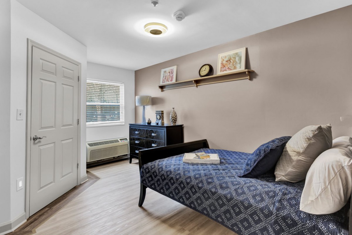 A tidy bedroom with minimalist decor, featuring a single bed, dresser, and a wall-mounted shelf.