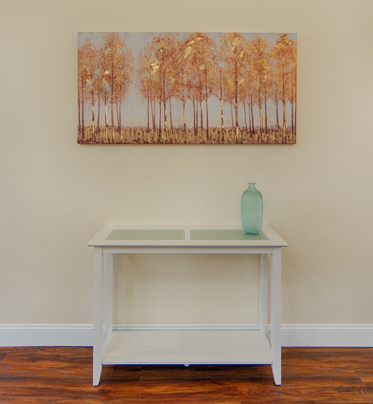 A minimalist interior setup featuring a white table with a light blue vase and a tree-themed painting on the wall.