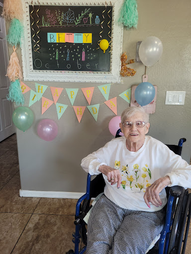 An elderly woman sits in a wheelchair surrounded by colorful decorations and balloons, celebrating a special occasion.