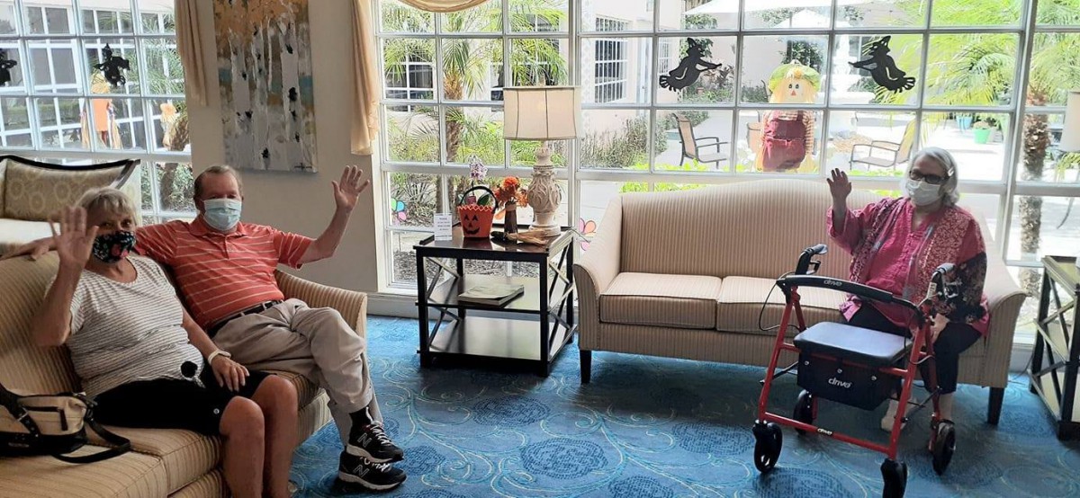 An elderly person sitting in a living room with a walker nearby, sunlight streaming through the window.