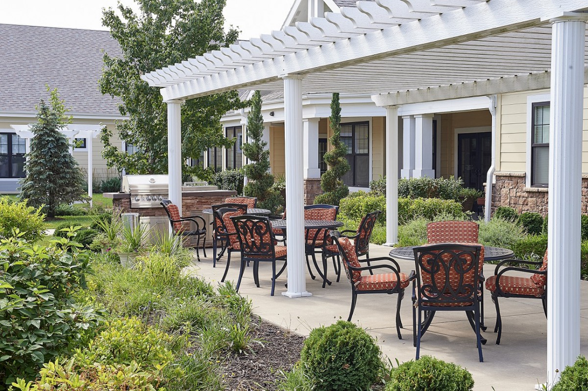 A patio area with outdoor seating under a white pergola surrounded by landscaped greenery.