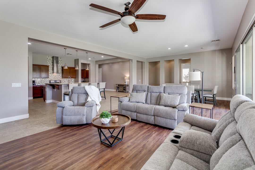 A modern living room with a reclining sofa set and ceiling fan, showcasing an open floor plan leading to the kitchen.