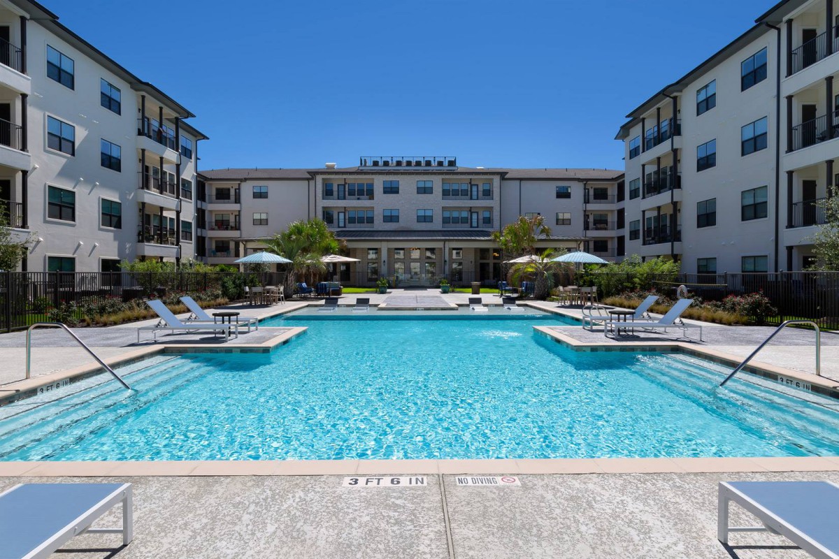 A modern apartment complex with a central swimming pool, surrounded by lounge chairs under a clear blue sky.