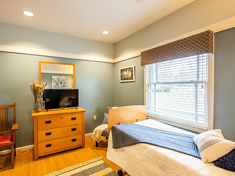 A cozy bedroom featuring a bed with a blue throw, a wooden dresser with a TV, and large windows with blinds.