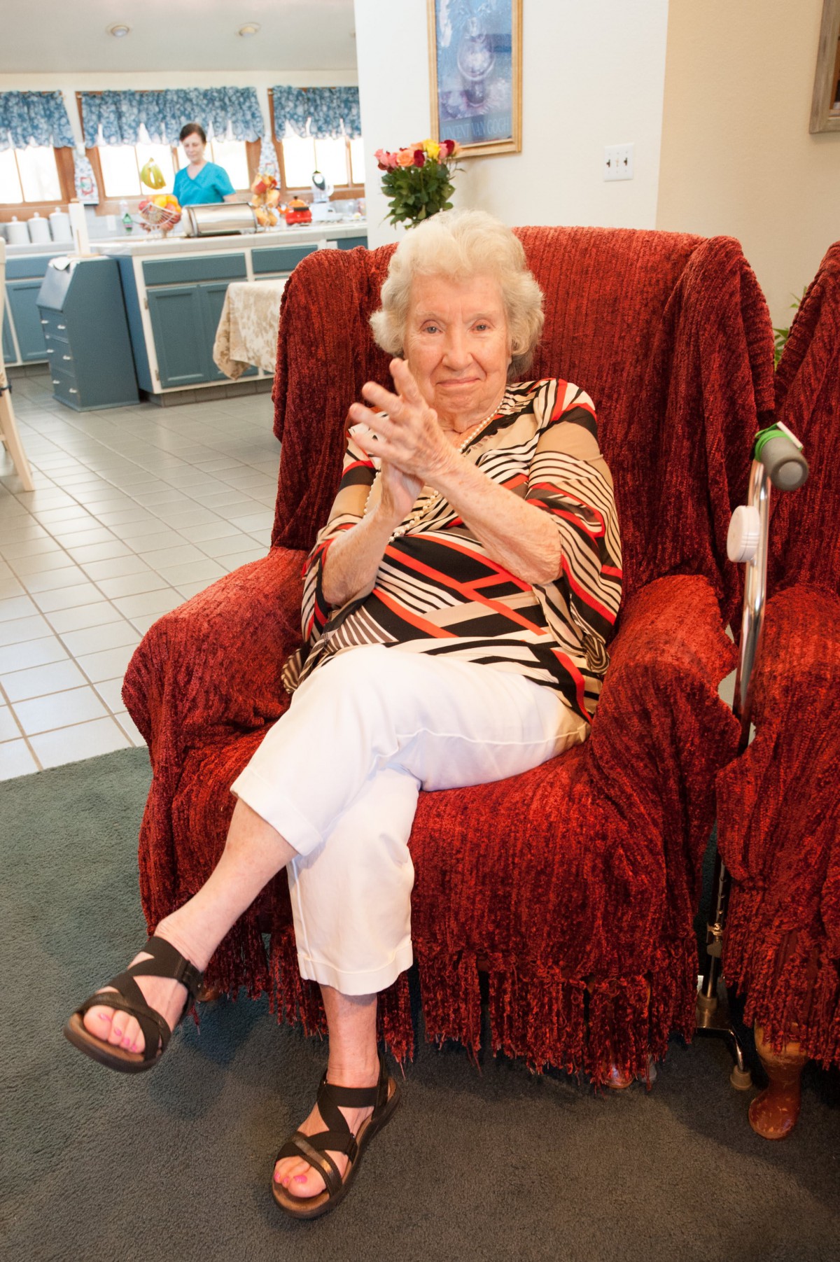 An elderly woman smiling while sitting on a red chair indoors.