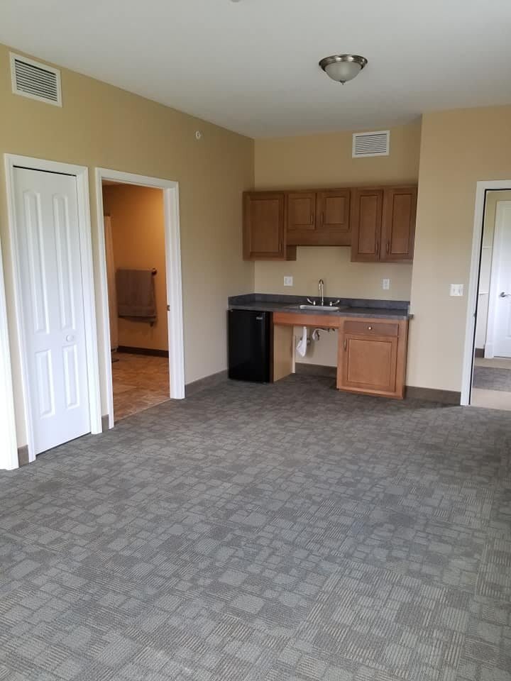 An empty room with a kitchenette, featuring cabinets and a sink, carpet flooring, and open doors.