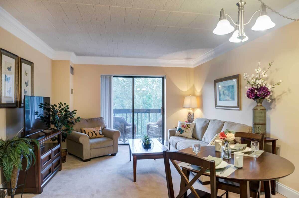A cozy living room with a sofa, armchair, and dining area, featuring a large window allowing natural light.