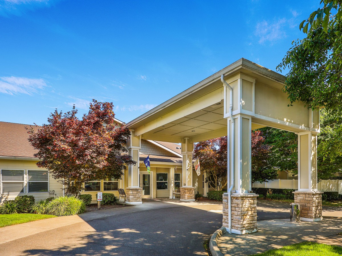 The image shows a building entrance with a covered driveway, landscaped with trees.