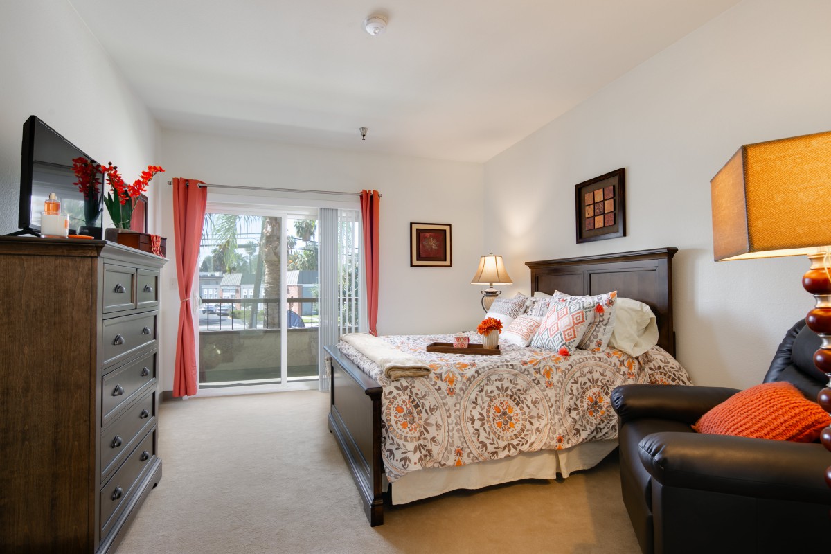A cozy bedroom with a large bed and colorful decor, featuring a window with red curtains and various pieces of artwork on the walls.