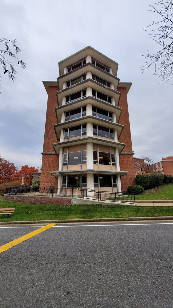 A modern brick building with multiple stories and large windows, situated on a green lawn by the street.
