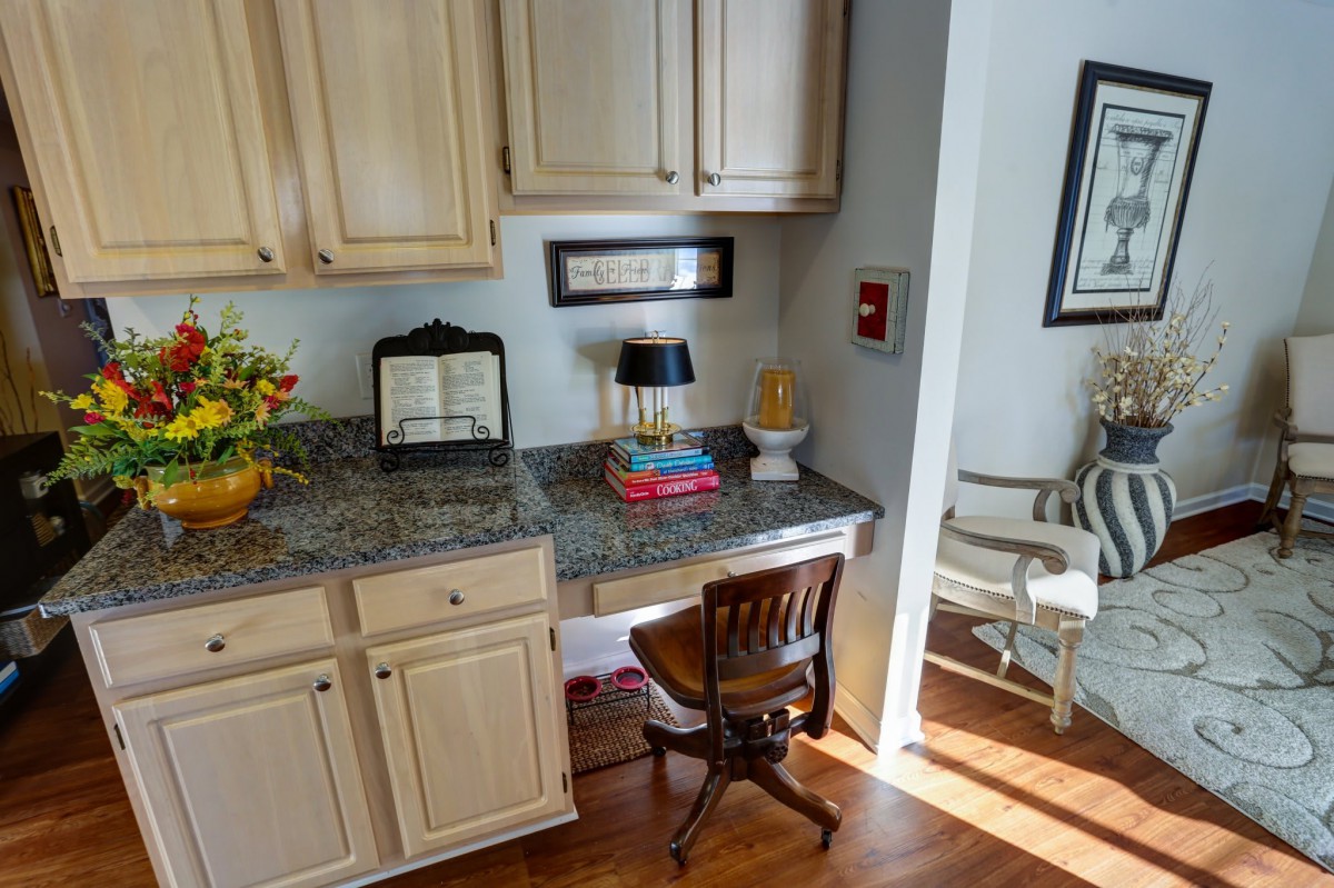 A cozy home office setup featuring a small desk with a wooden chair, surrounded by cabinetry and decor, including a lamp and flowers.