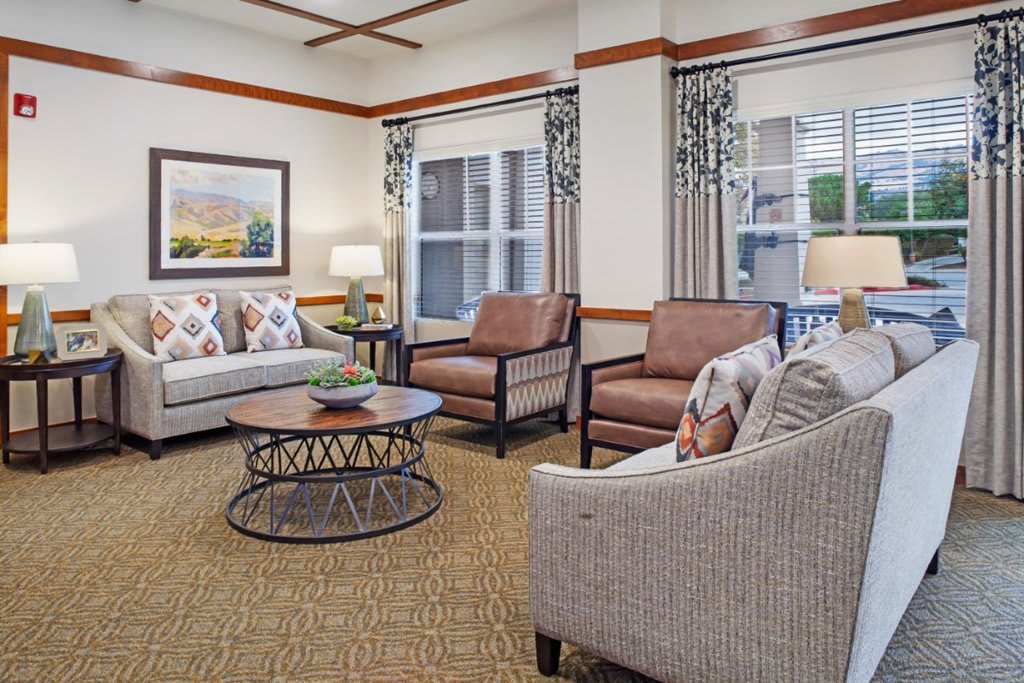 A cozy living room with comfortable sofas and armchairs arranged around a circular coffee table, featuring elegant decor and large windows with patterned curtains.