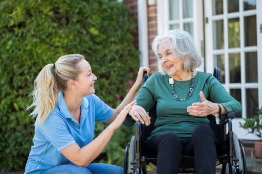 A caregiver interacts warmly with an elderly woman in a wheelchair, providing companionship and support in an outdoor setting.