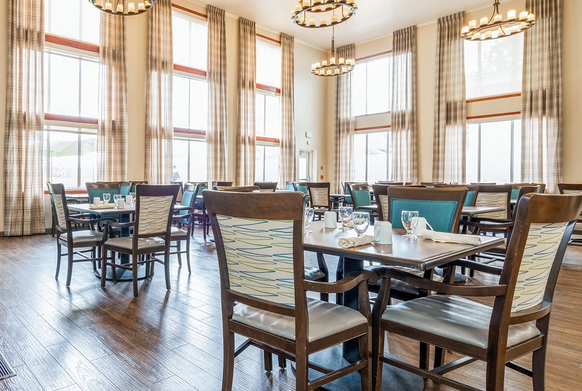 A spacious and well-lit dining room with neatly arranged tables and chairs, featuring large windows and elegant chandeliers.