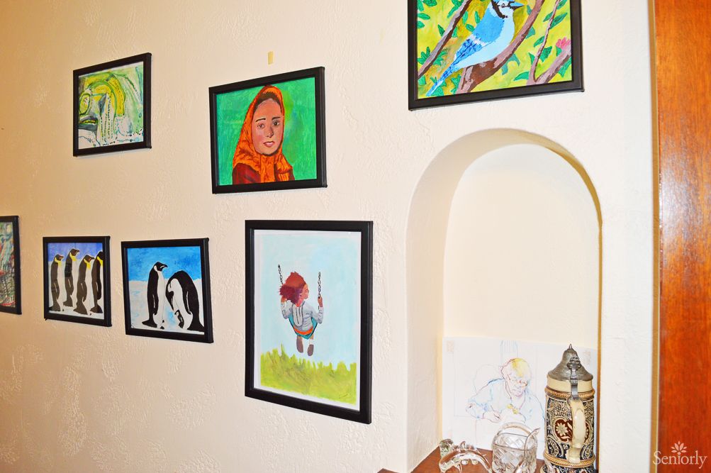 A collection of framed paintings displayed on a wall, featuring birds, animals, and portraits in a cozy interior setting.