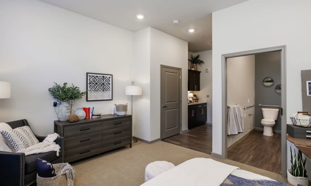 A modern bedroom interior with a dresser and lamp, offering a view into an adjacent kitchen.