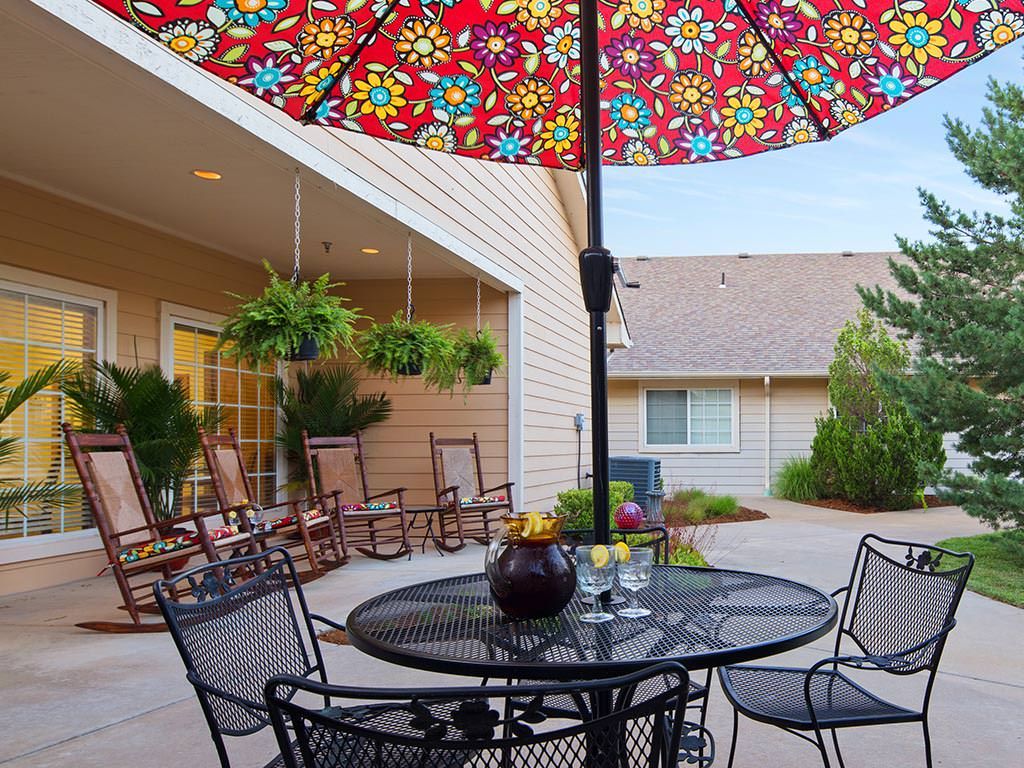 A cozy outdoor patio with a colorful umbrella, rocking chairs, and a table set for relaxation.