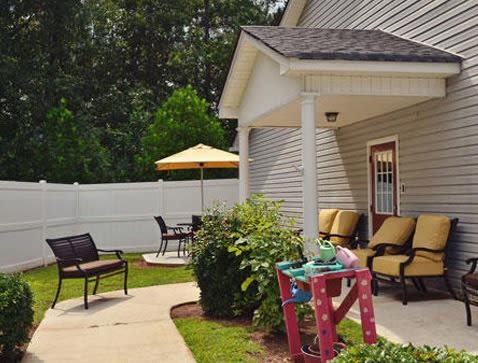 A cozy patio area with outdoor seating, including chairs and a table under an umbrella, surrounded by greenery and a white fence.