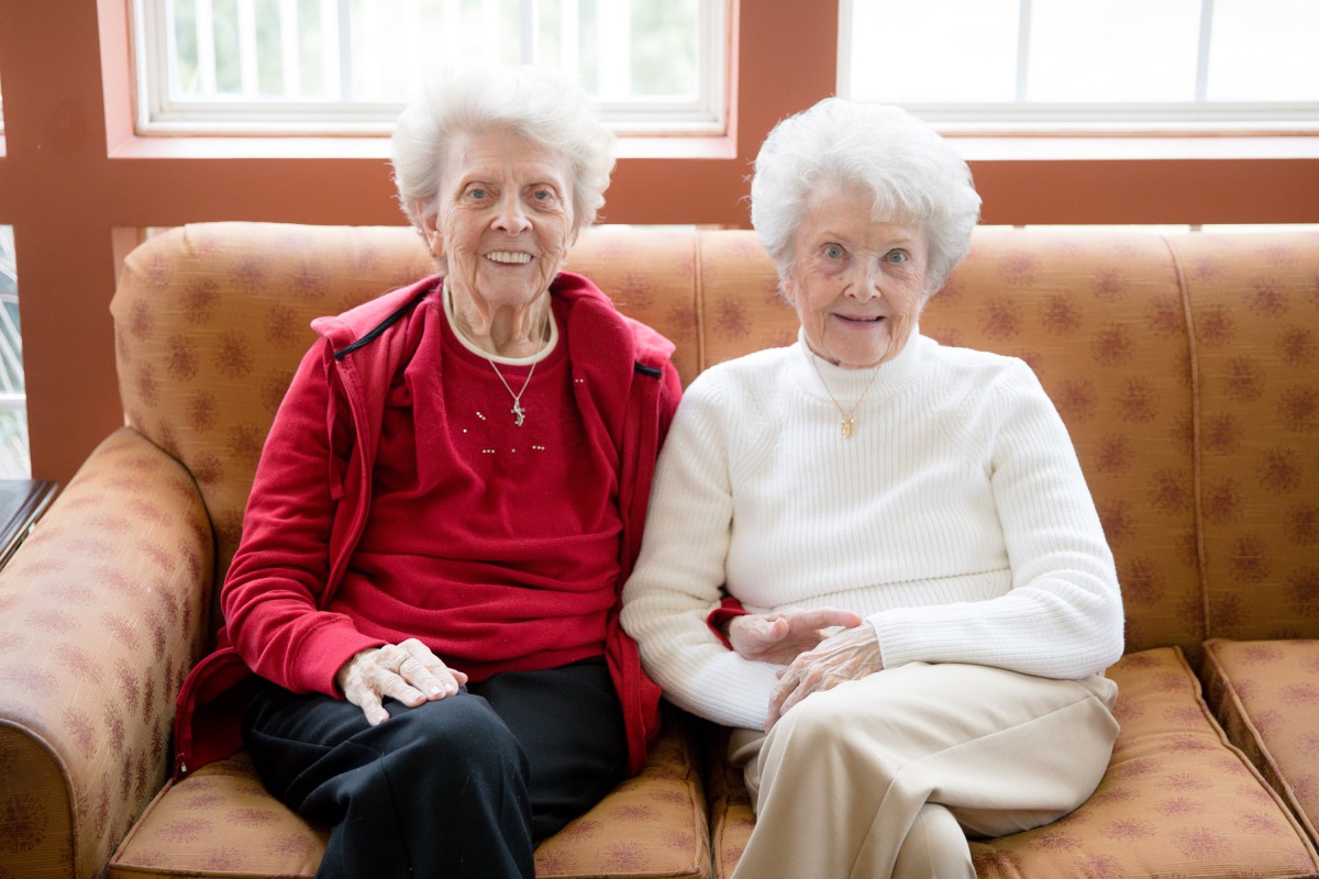 Two elderly women sitting together on a couch, smiling.