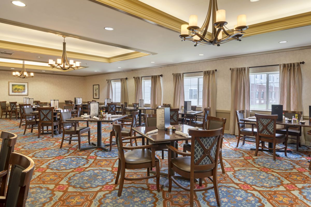 A spacious dining area with elegant chandeliers, neatly arranged tables and chairs, and vibrant carpet.
