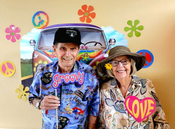 A cheerful couple dressed in fun retro outfits stands in front of a colorful vintage car, embodying the spirit of the 1960s.