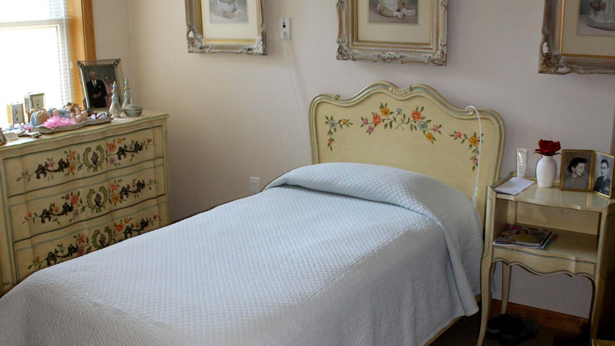 A classic bedroom featuring a single bed with a floral-patterned headboard, accompanied by a dresser and framed artwork.