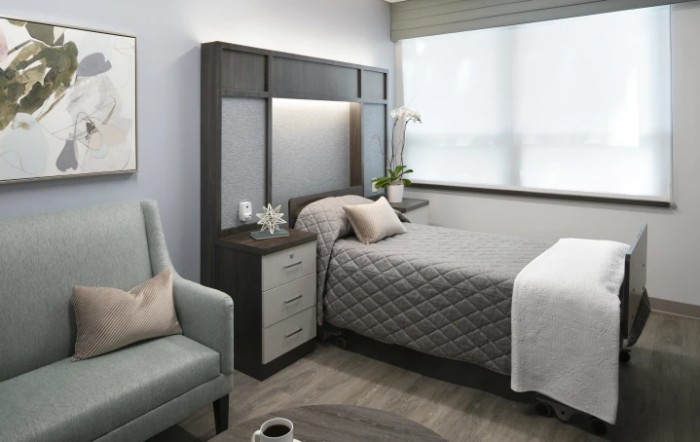 A modern bedroom with a gray quilted bed, bedside table, and an armchair near a large window.