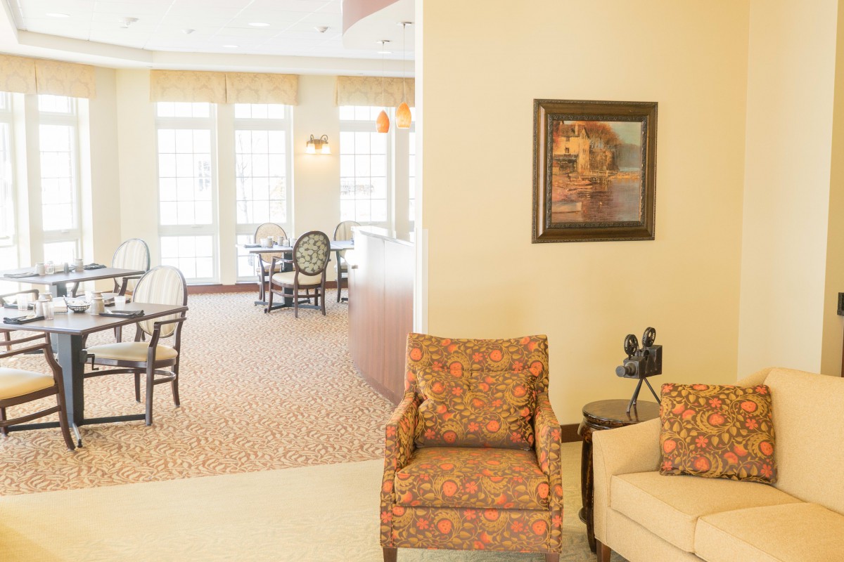 A bright and inviting seating area with comfortable chairs and artwork on the wall, leading to a dining space with multiple windows.
