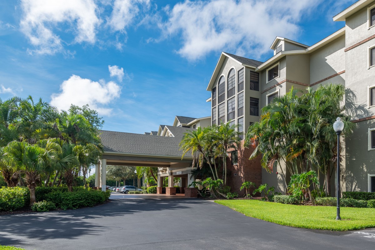 A hotel with a welcoming entrance surrounded by palm trees and greenery under a bright blue sky.