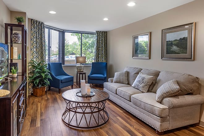 A cozy living room with a comfortable sofa, blue chairs, and a round coffee table, featuring hardwood floors and large windows.
