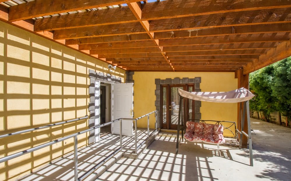 A sunny patio with a wooden pergola casting shadows, featuring an outdoor swing and yellow walls.