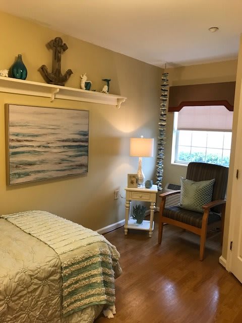 A cozy bedroom with nautical-themed decor, featuring a bed, a lamp on a nightstand, and a comfortable chair by the window.
