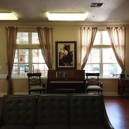 A cozy living room with a piano placed between two windows, adorned with curtains and an artwork above the piano.