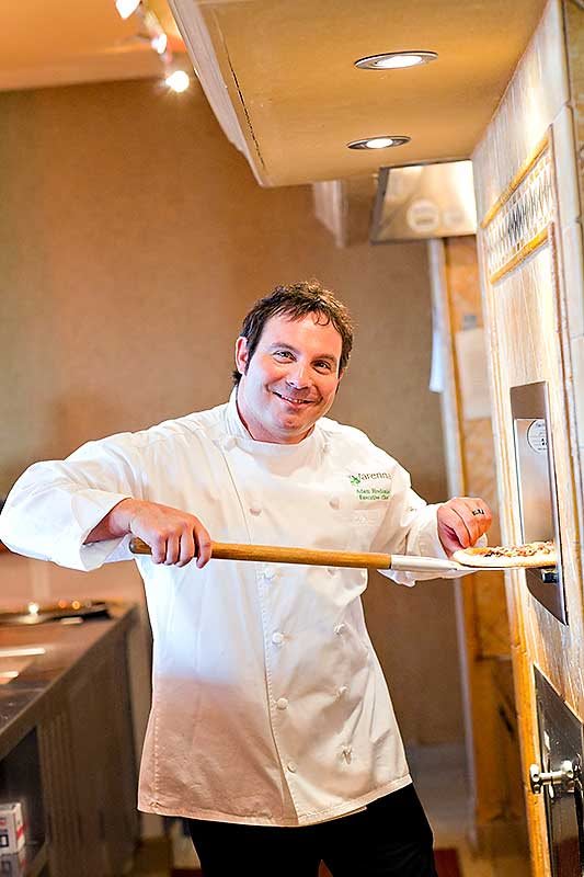 A chef in a white uniform smiling while preparing pizza in a kitchen.