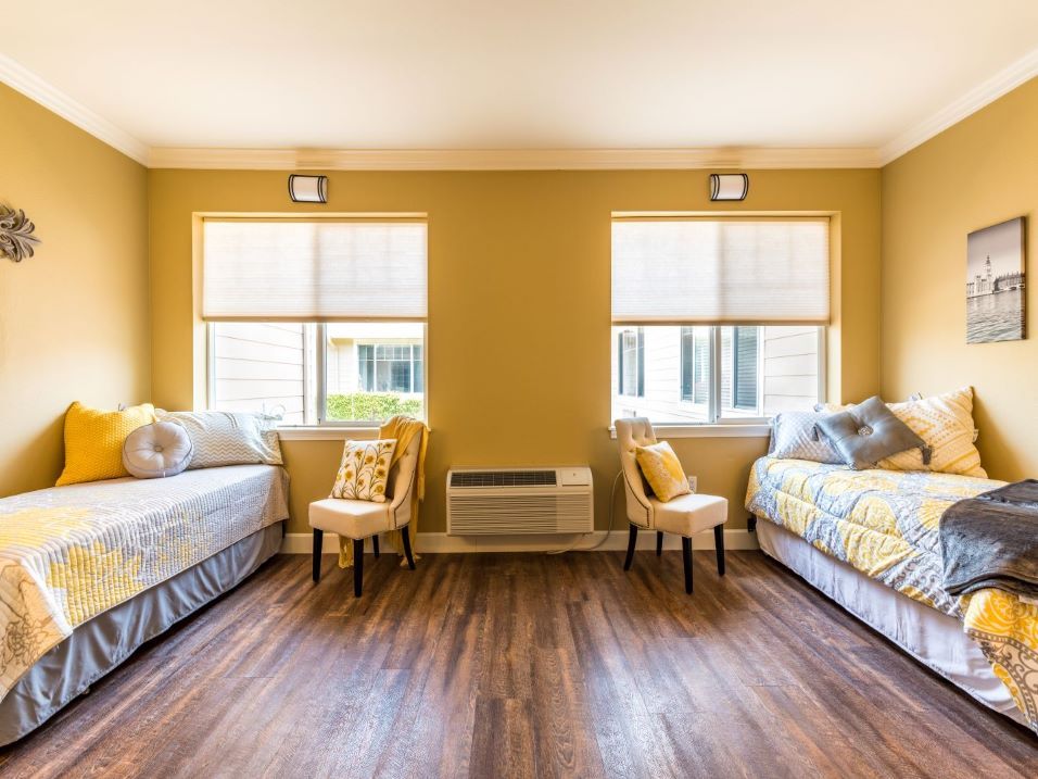 A bright bedroom featuring twin beds with yellow decor, wooden flooring, and two windows.