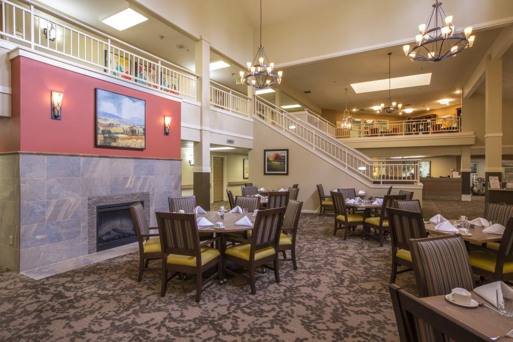 A spacious dining area with elegant chandeliers, neatly arranged tables and chairs, a carpeted floor, and a cozy fireplace.