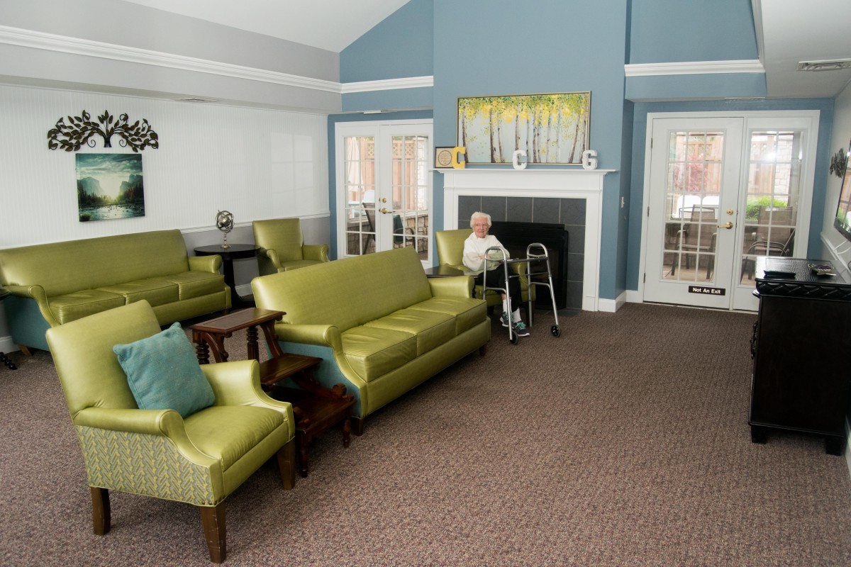 A senior person using a walker sits in a furnished living room with couches, chairs, and a fireplace.