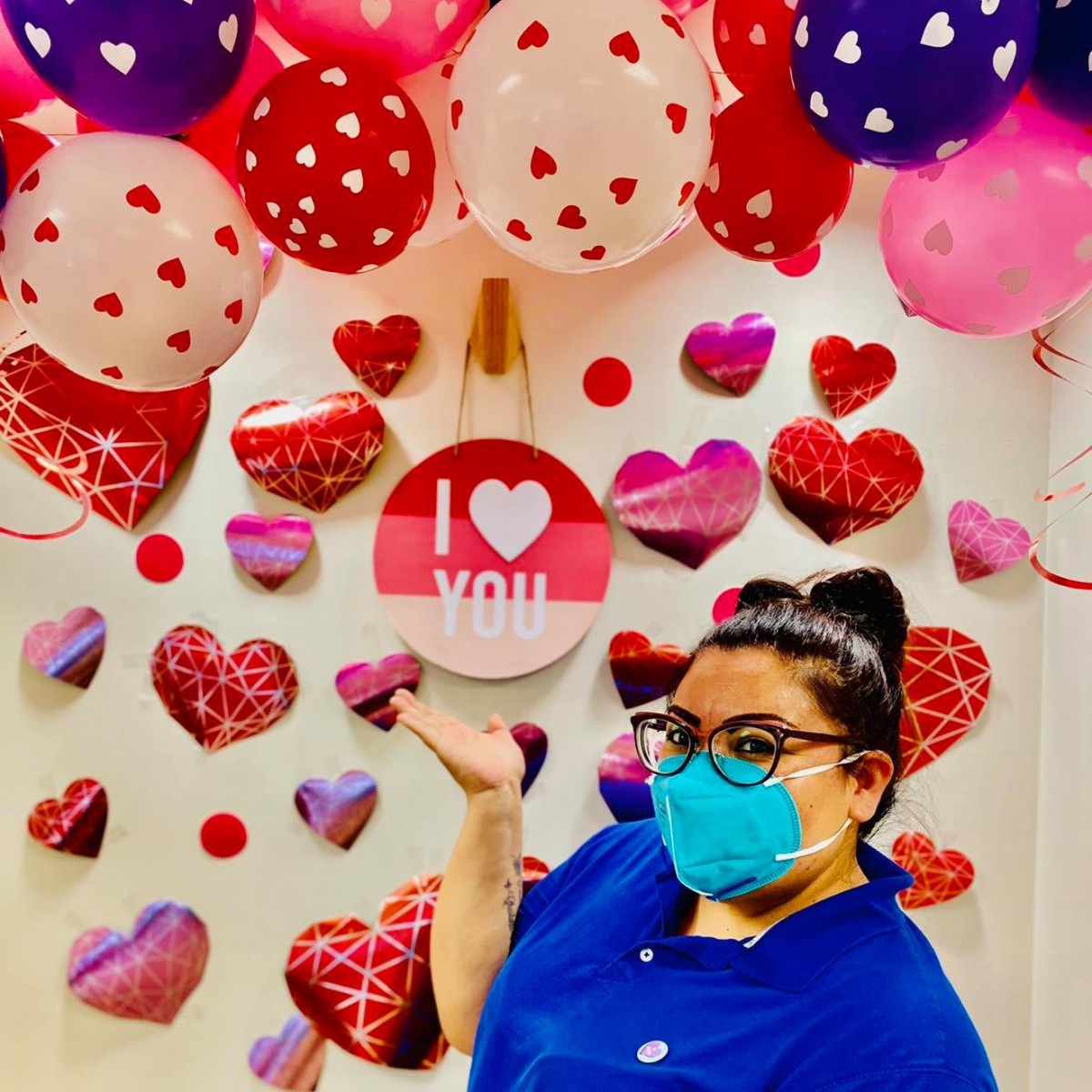 A person in a blue shirt and mask stands in front of a festive background of heart-shaped decorations and balloons, celebrating Valentine's Day.