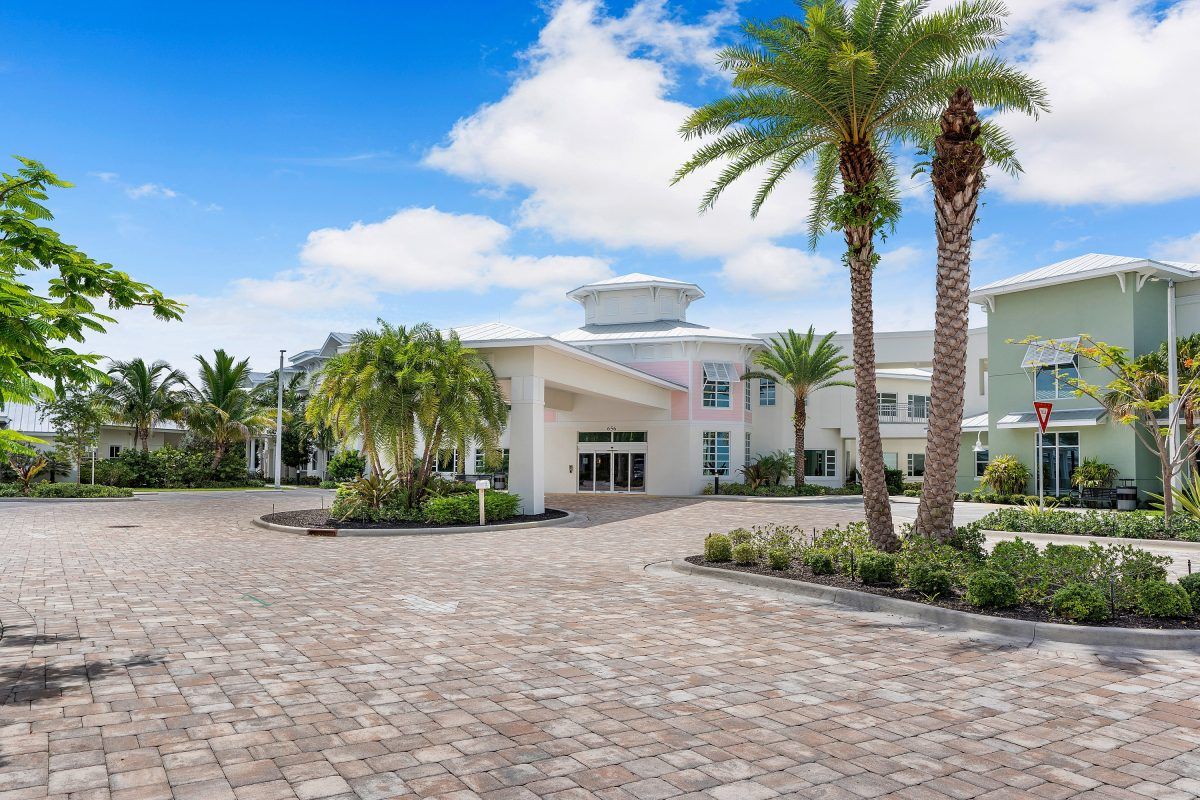 A white modern building surrounded by palm trees and a paved driveway under a partly cloudy sky.