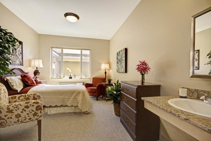 A cozy bedroom with neutral tones, featuring a bed, dresser, armchair, lamps, and a large window allowing natural light.