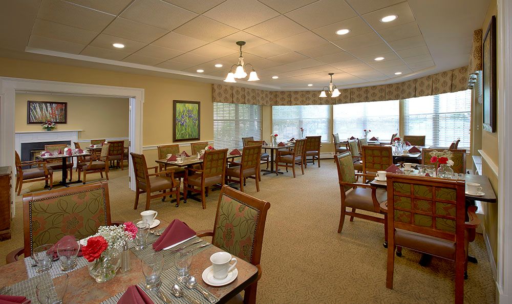 A cozy dining room with neatly arranged tables and chairs, featuring place settings and ample natural light from large windows.