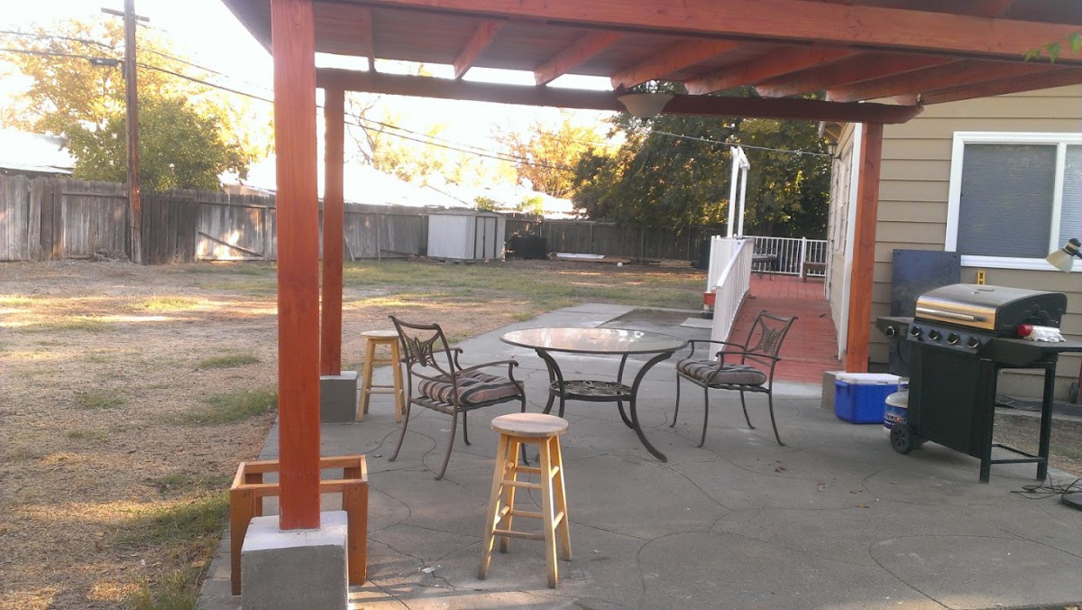 An outdoor patio area with a table and chairs under a pergola, set in a backyard.